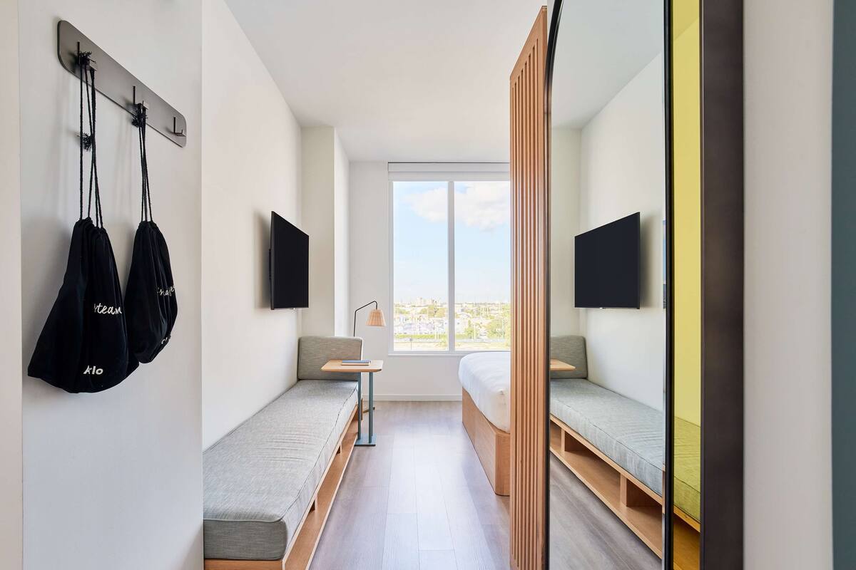 A modern, compact room with a bed, a bench, and a TV mounted on the wall.