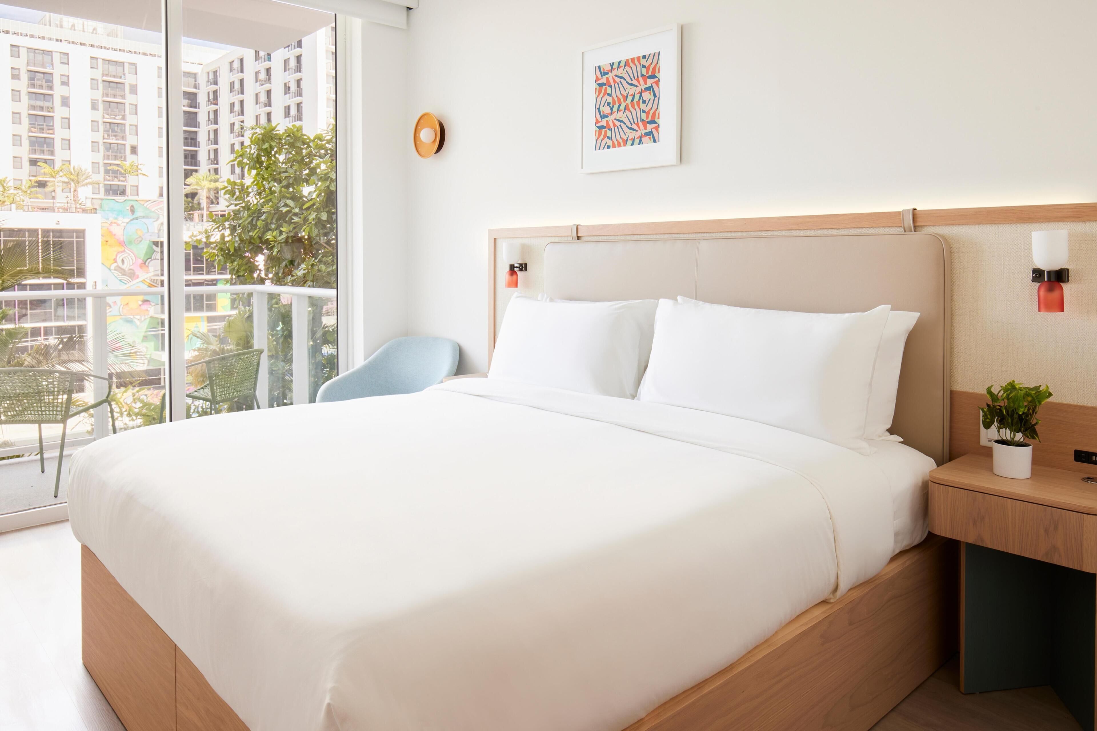 Quarto de hotel com uma cama grande, uma mesa de cabeceira com abajur, uma cadeira, uma planta em vaso e uma janela com vista para prédios e vegetação.