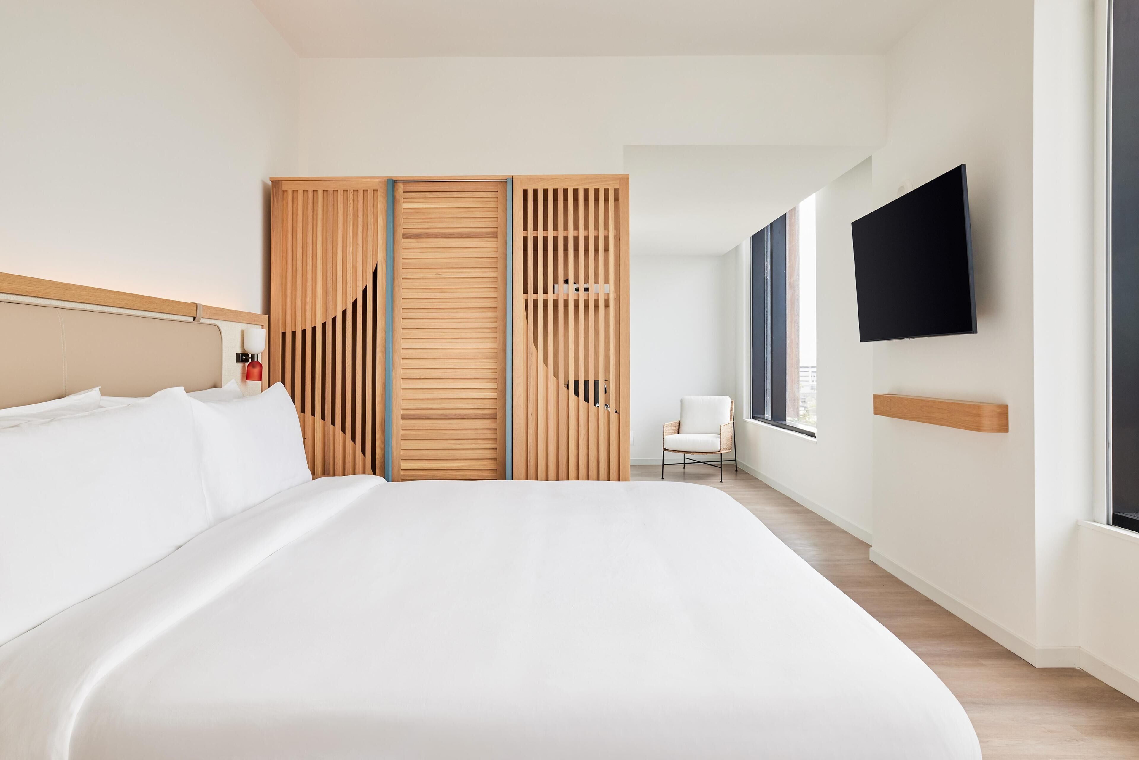 A modern hotel room with a large bed, a wooden wardrobe, a white chair, and a flat-screen TV mounted on the wall.