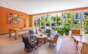 A pool table with surrounding wicker chairs and a table, large windows with a view of greenery, and a colorful abstract painting on the wall. - Arlo Wynwood Miami (Miami)