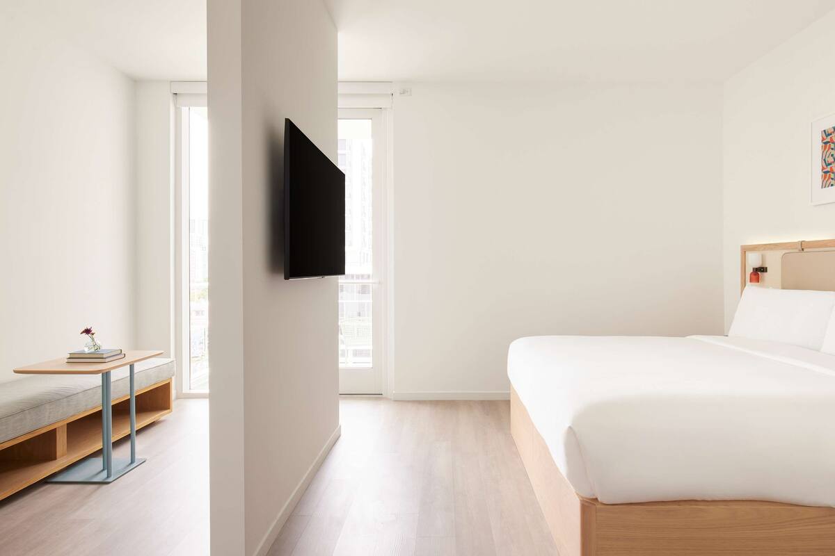 A modern hotel room with a bed, a TV mounted on the wall, a small table with a book and vase, and a window with a view of the city.