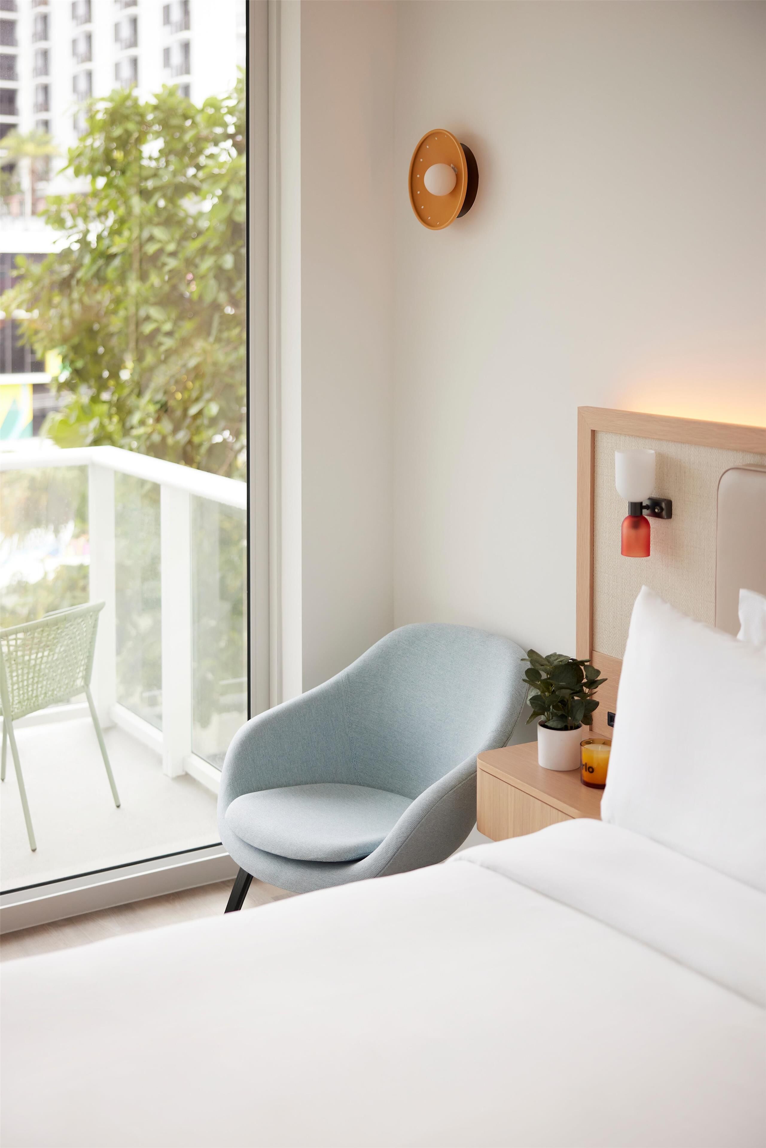 A hotel room with a bed, a chair, a bedside table with a lamp, a small plant, and a view of greenery outside.