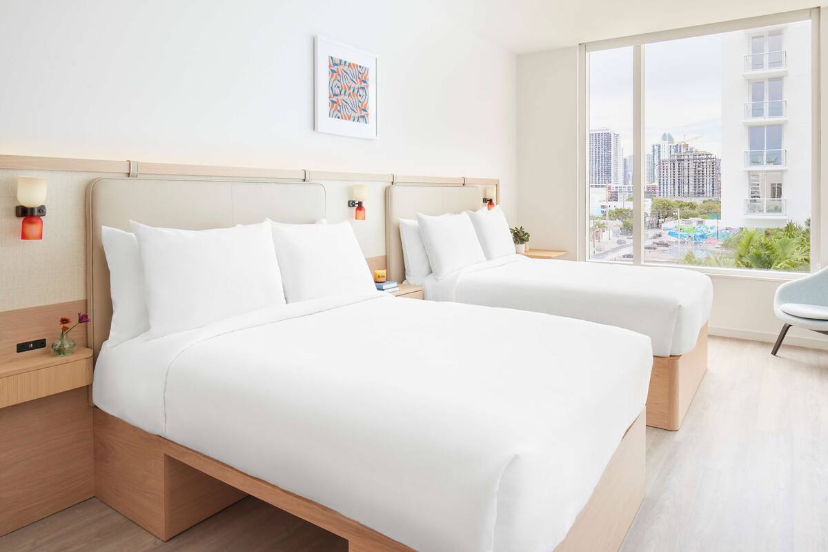 A hotel room with two beds, a large window with a city view, a small table with a vase, and a wall-mounted light fixture.