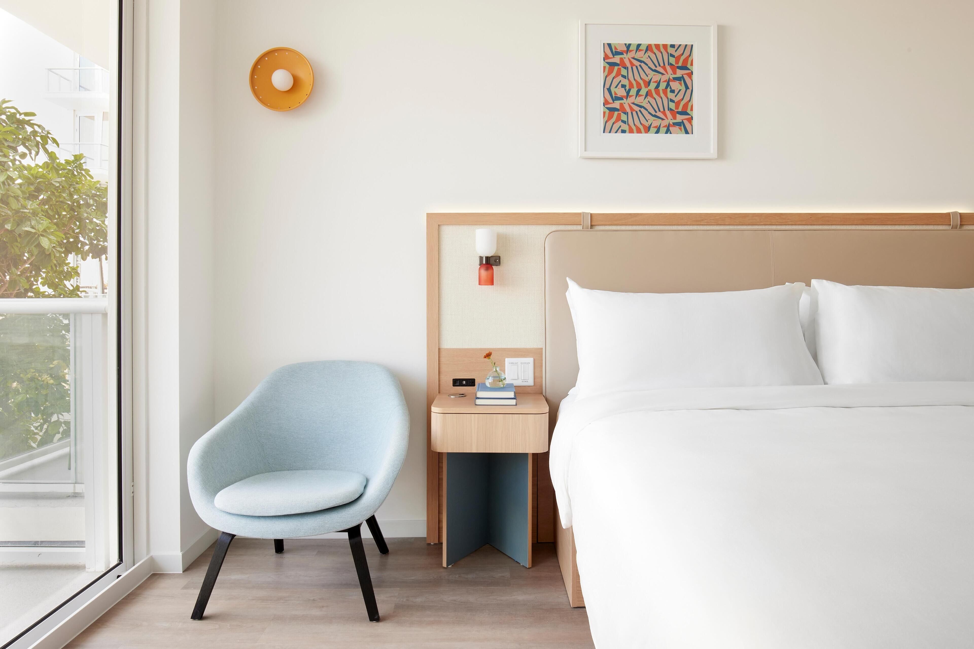 Camera d'albergo con un letto, una sedia, un comodino, una lampada a parete e un quadro astratto incorniciato.