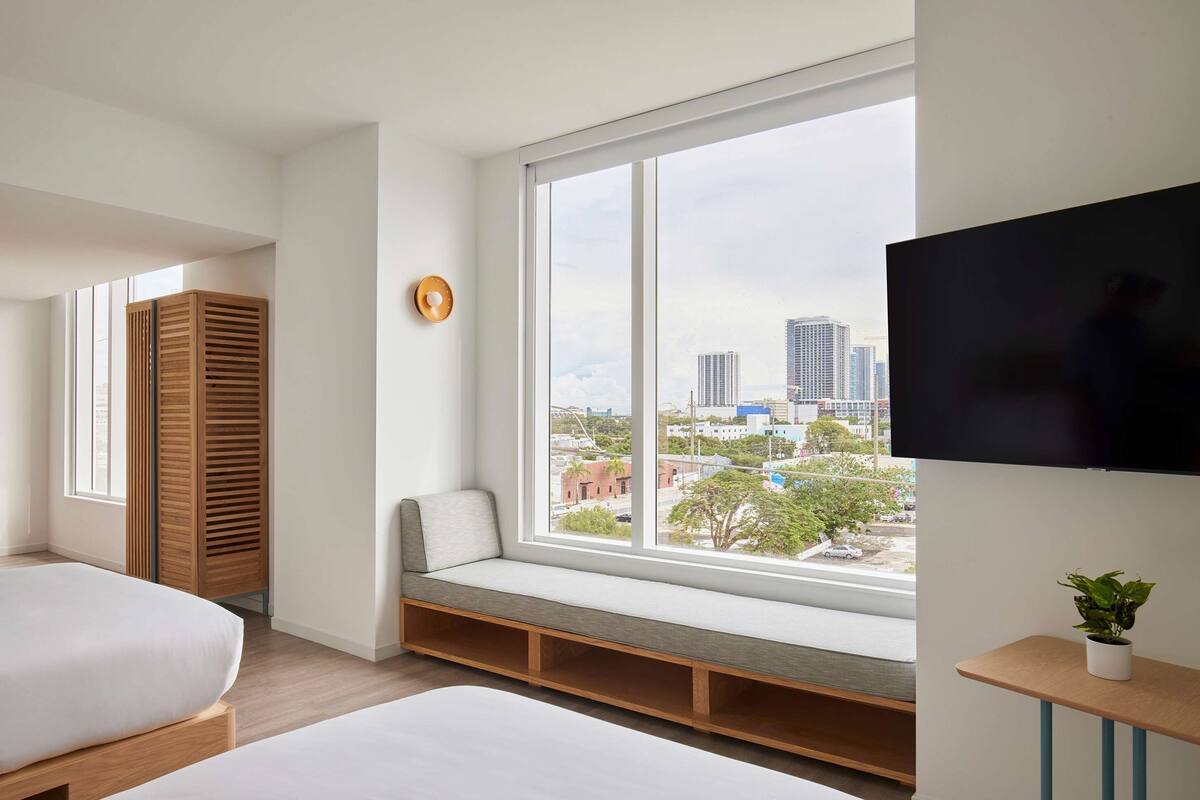 A modern hotel room with a large window, a bed, a seating area, and a TV.