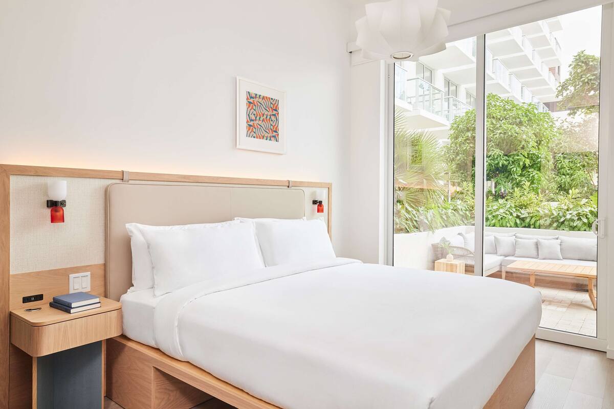 A modern hotel room with a large bed, a bedside table with a book, a wall-mounted abstract art piece, and a view of a balcony with greenery.
