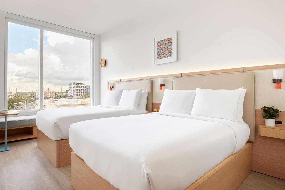 A hotel room with two beds, large windows, a view of the city, and a small table.