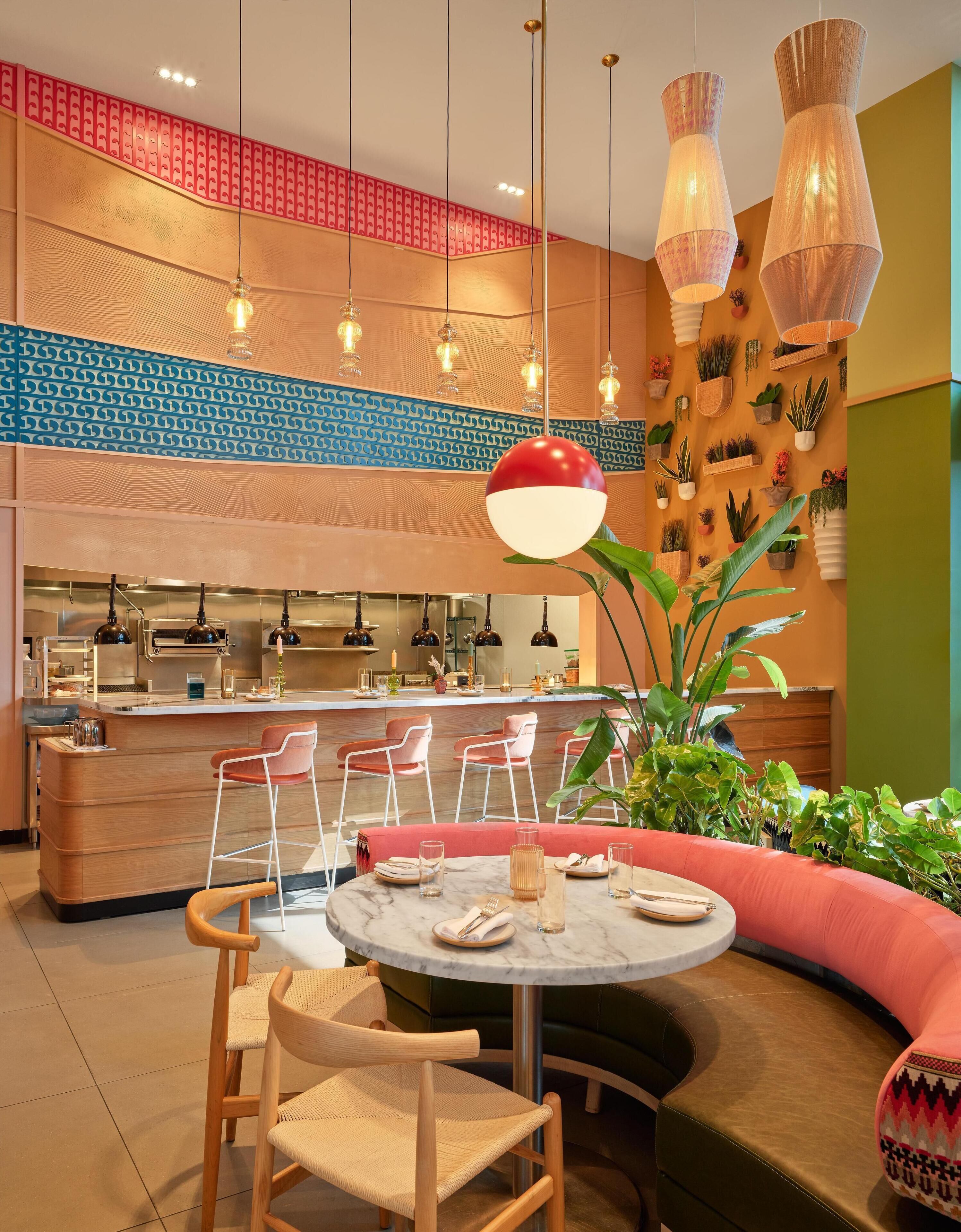 A modern restaurant with a central dining area featuring a round table and chairs, a kitchen in the background, and decorative plants and wall art.
