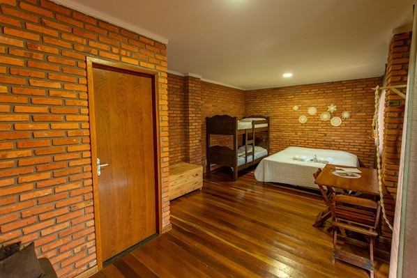 Una habitación con pared de ladrillo, puerta de madera, literas, una mesa pequeña con sillas y un banco de madera.