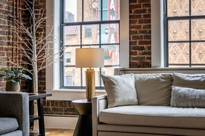 Ein gemütliches Wohnzimmer mit einer Couch, einer Lampe, einer Pflanze und Blick auf die Stadt durch große Fenster.