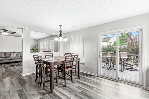 Un coin repas avec une table et des chaises en bois, un ventilateur de plafond et une vue sur une terrasse extérieure, accessible par une porte-fenêtre coulissante.