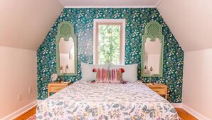 A bedroom with a sloped ceiling, floral wallpaper, a bed with a floral bedspread, two side tables with lamps, and a window with a view of greenery. - The Boho Loft: colorful, cozy, comfortable 1BR (Hot Springs)
