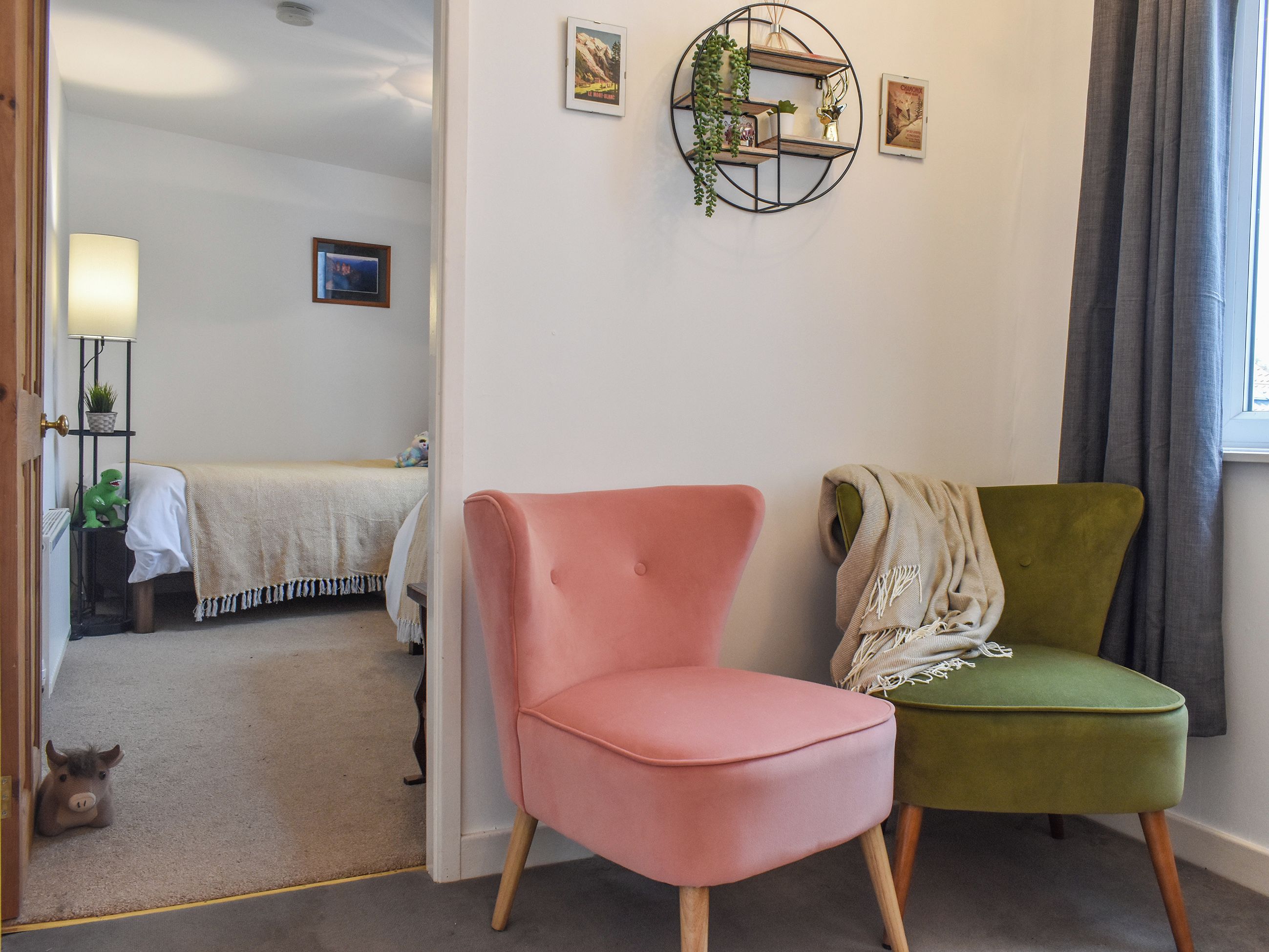 Une pièce comprenant un lit, un fauteuil rose, un fauteuil vert, une étagère fixée au mur et une fenêtre avec des rideaux.