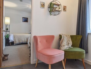 Una habitación con una cama, una silla rosa, una silla verde, un estante montado en la pared y un ventanal con cortinas.