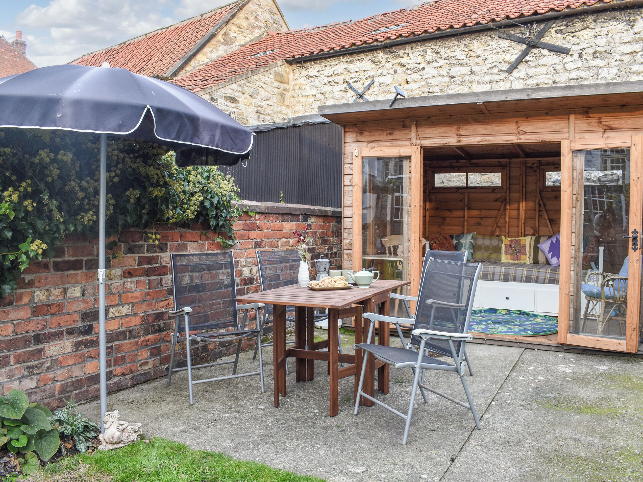 A patio area with a wooden table and chairs, a blue umbrella, and a brick wall with a stone building in the background.