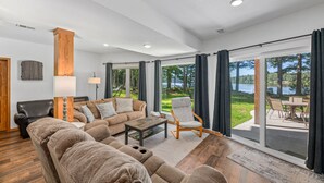 A spacious living room with a large sofa, a wooden coffee table, and a chair. There is a view of a patio with outdoor furniture and a body of water.