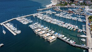 Une vue aérienne d’une marina où sont amarrés de nombreux bateaux, avec une jetée qui s’avance dans l’eau et une ville côtière en arrière-plan.