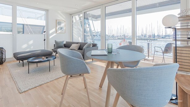 Un salon moderne doté d’une grande fenêtre donnant sur une marina. On y trouve deux canapés, une table basse et une table à manger avec des chaises.