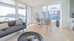 Un salon moderne comprenant un canapé, une table à manger et des chaises, de grandes fenêtres offrant une vue sur l’eau, ainsi qu’un coin cuisine.