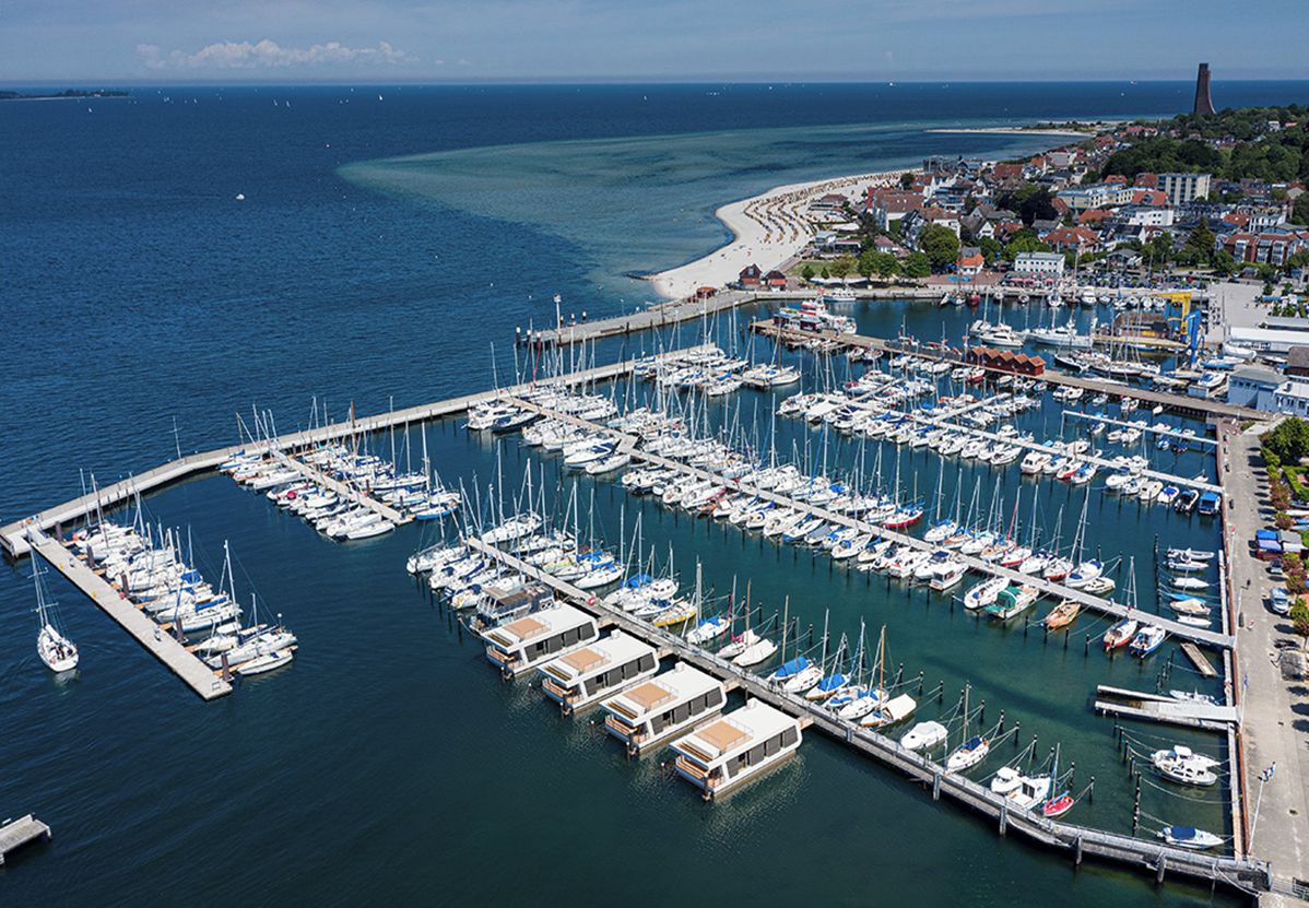 Vista aèria d'una marina amb nombrosos vaixells amarrats, un moll que s'endinsa a l'aigua i una població costanera al fons.