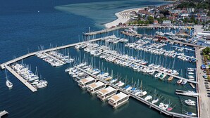 Eine Luftaufnahme eines Yachthafens mit zahlreichen dort liegenden Booten, einer Pier, die sich ins Wasser erstreckt, und einer Küstenstadt im Hintergrund.