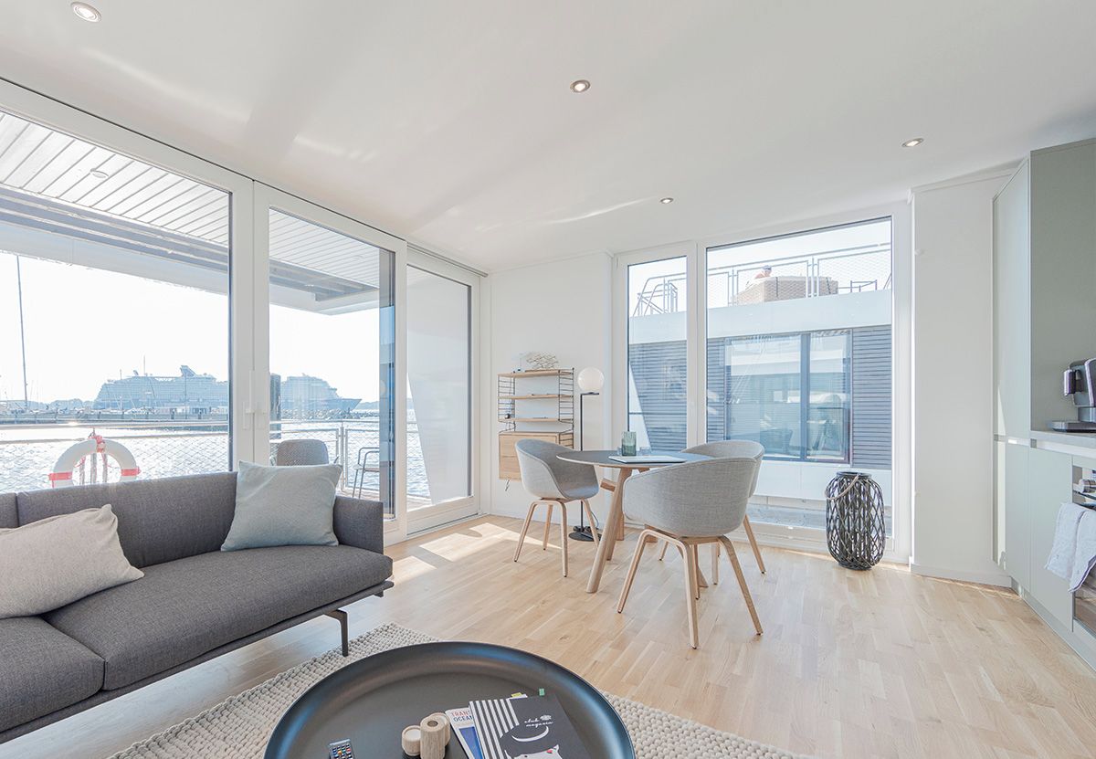 Una sala de estar moderna con sofá, mesa de comedor, sillas, ventanas amplias con vista al agua y una zona de cocina.