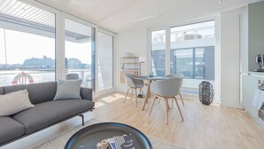 Ein modernes Wohnzimmer mit Sofa, Esstisch, Stühlen, großen Fenstern mit Blick auf das Wasser und einer Küchenzeile.
