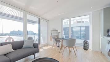 Una sala de estar moderna con sofá, mesa de comedor, sillas, ventanas amplias con vista al agua y una zona de cocina.