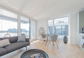 A modern living room with a sofa, dining table, and chairs, large windows with a view of the water, and a kitchen area. - Houseboat wake FH-LA 03 (Laboe)