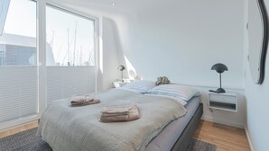 Une chambre moderne avec un grand lit, des tables de chevet et une vue sur les toits de la ville grâce à de grandes fenêtres du sol au plafond.