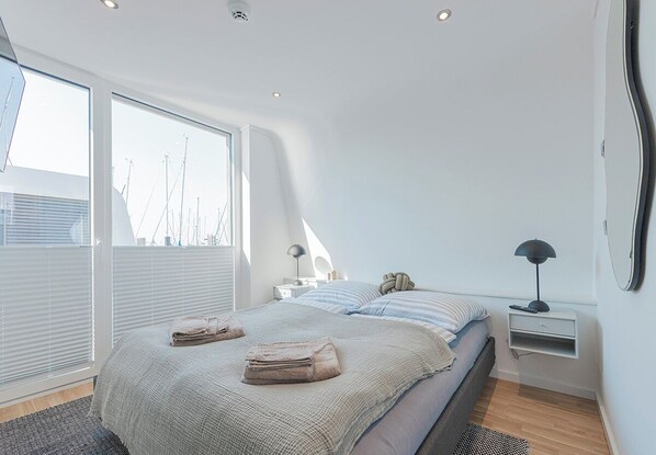 A modern bedroom with a large bed, bedside tables, and a view of the city skyline through floor-to-ceiling windows. - Houseboat wake FH-LA 03 (Laboe)