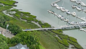 An aerial view of a marina with multiple boats docked, a bridge connecting to a residential area, and lush greenery surrounding the water.