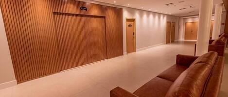 Pasillo de hotel con pared de paneles de madera, una zona de recepción y un área de descanso con sofás marrones.