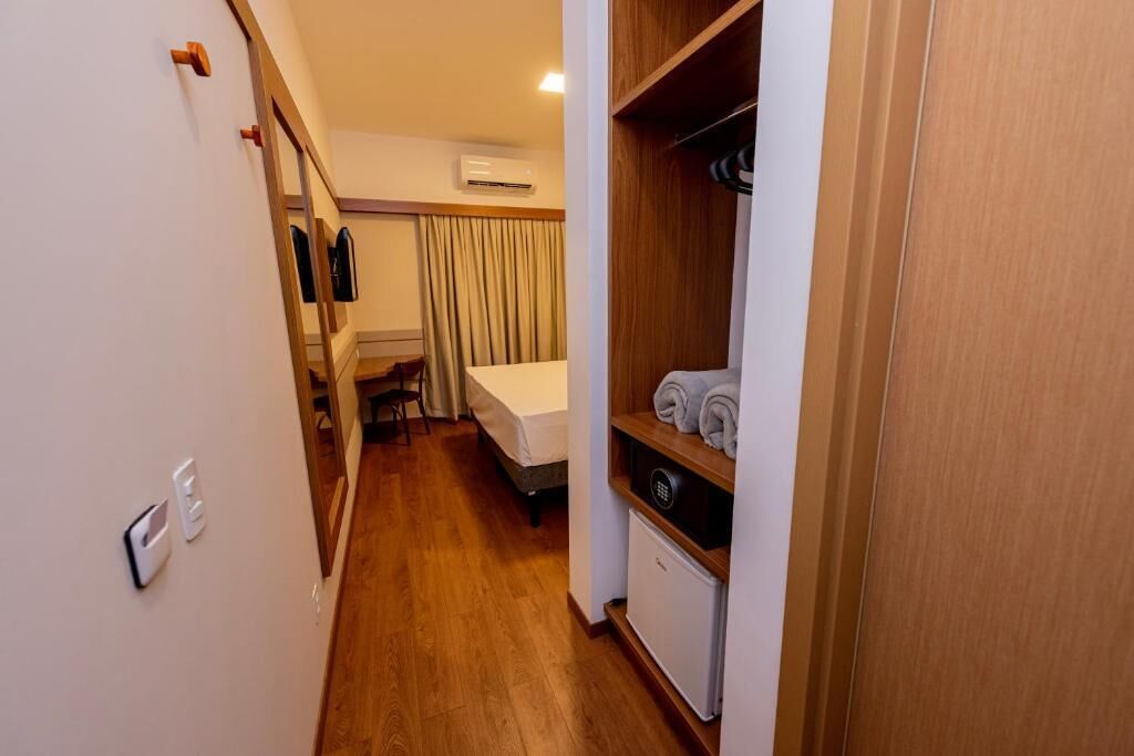 A narrow hallway leading to a bedroom with a bed, a small table, and a chair. There is a wooden wardrobe with a shelf holding towels. An air conditioning unit is mounted on the wall above the bed.
