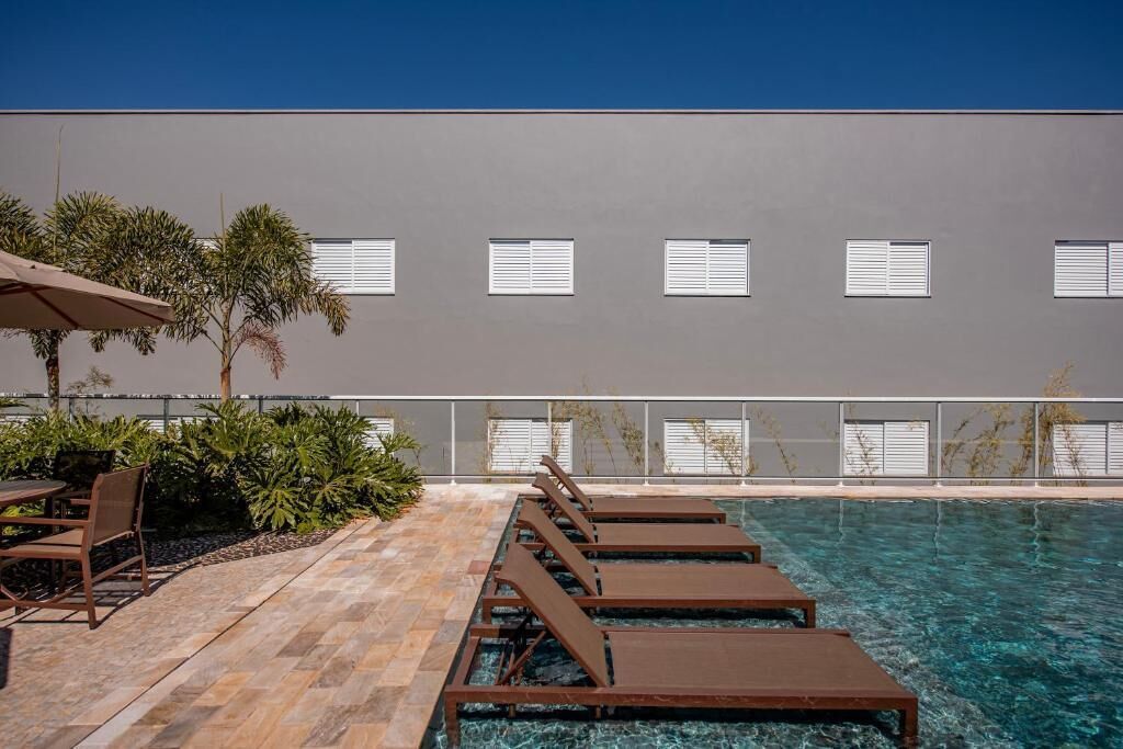 Zona de piscina con sillones de descanso y una mesa, cielo azul despejado y un edificio con ventanas.