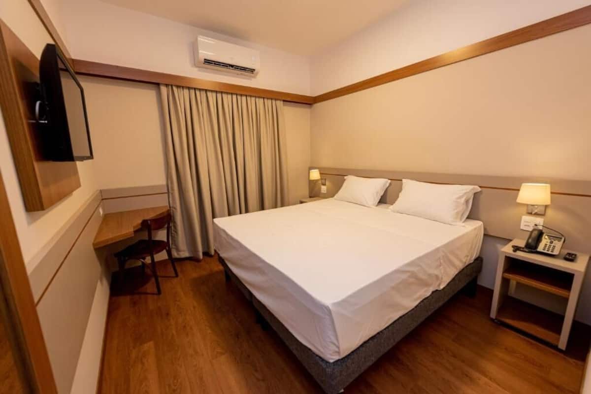 Habitación de hotel con cama, escritorio, silla, televisor y mesita de noche con lámpara.