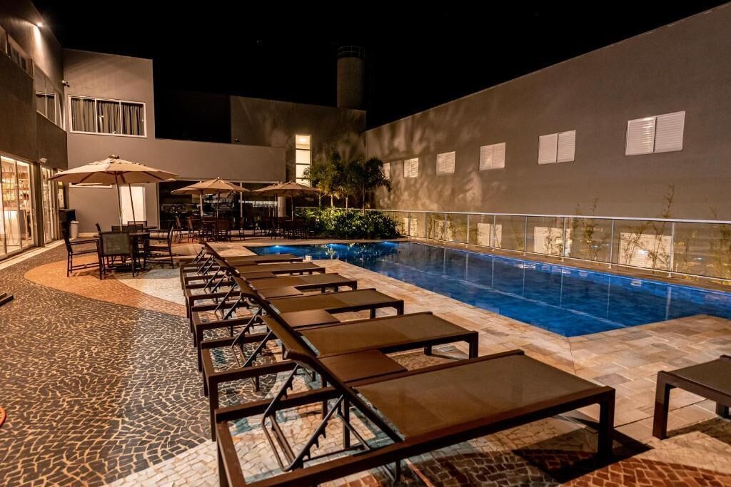 A pool area with lounge chairs and umbrellas, surrounded by a building with large windows and a tiled floor.