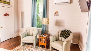 A room with two armchairs, a small wooden side table, a patterned rug, a window with blue curtains, an air conditioning unit, and a wall-mounted TV.