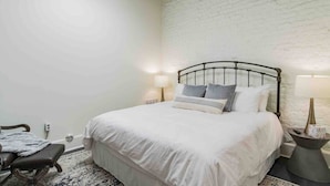 A bedroom with a white brick wall, a metal bed frame, a white bedspread, two bedside lamps, and a small table with a lamp. - Fabulous Loft w/ King Suite Great Location! (Birmingham)