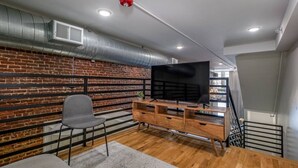 A modern living room with a brick wall, a flat-screen TV on a wooden stand, a grey chair, and a staircase with a metal railing. - Fabulous Loft w/ King Suite Great Location! (Birmingham)