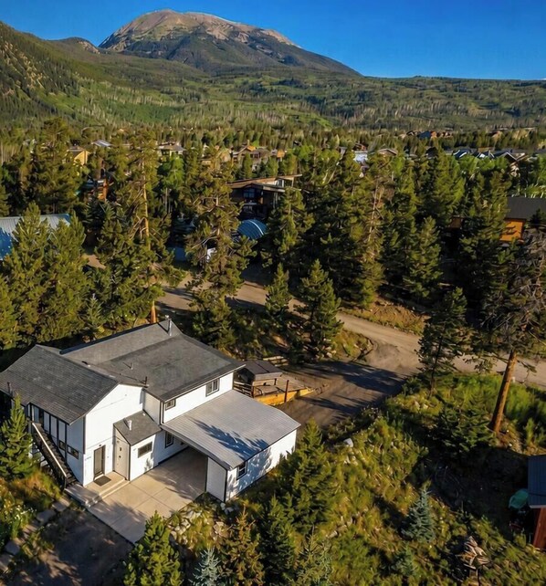 Una casa moderna de dos pisos con un amplio garaje, rodeada de un bosque frondoso y una montaña al fondo.