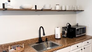 A kitchen with white cabinets, a granite countertop, a stainless steel sink, a microwave, a coffee maker, and a shelf with dishes and cups.
