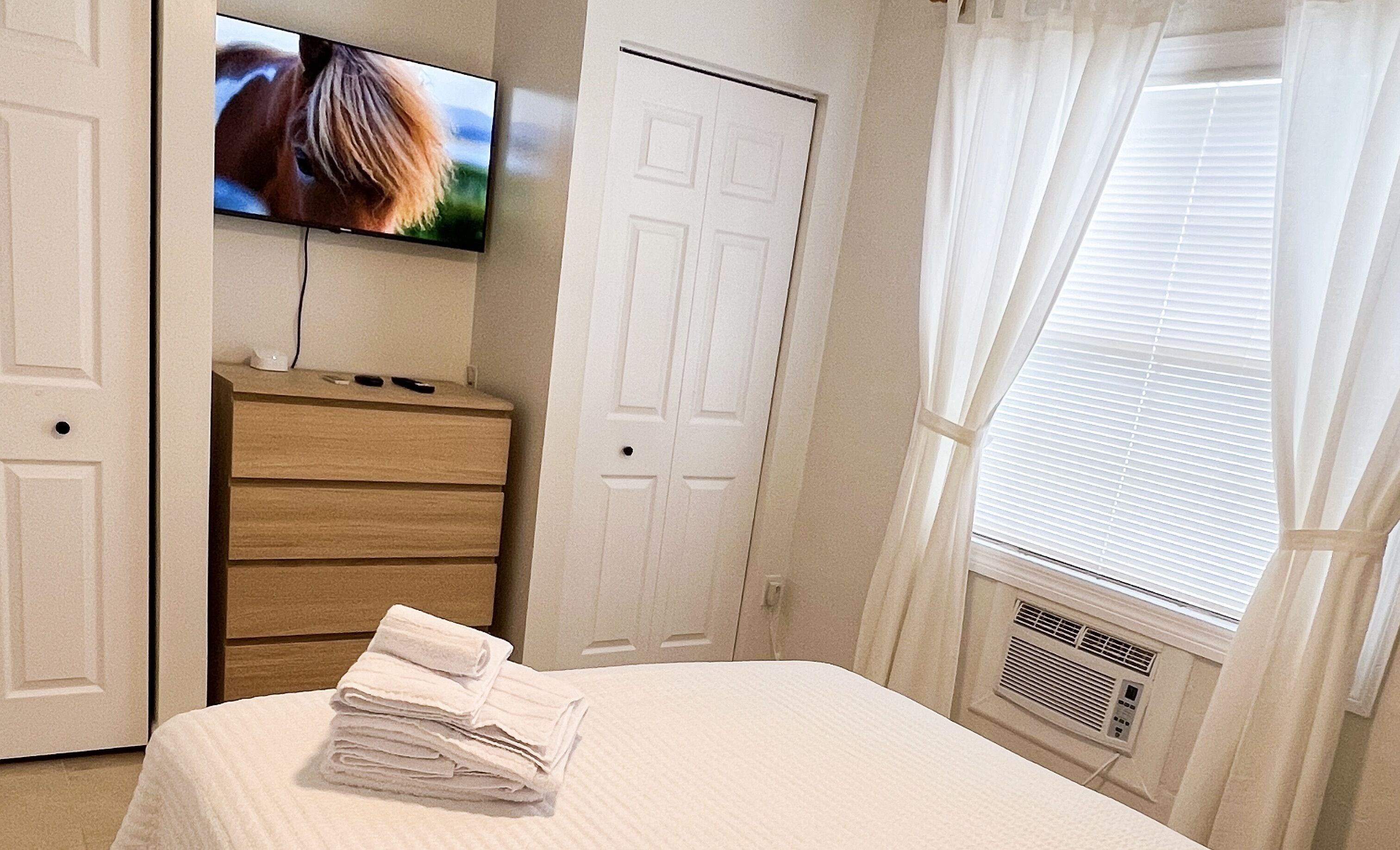 A bedroom with a bed, a dresser, a television on the wall, and a window with curtains.