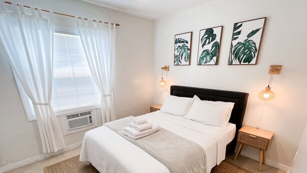 A neatly made bed with white linens, a woven rug, and a window with white curtains.