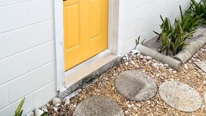 A yellow door with a window, white siding, and a small garden with pebbles and plants.