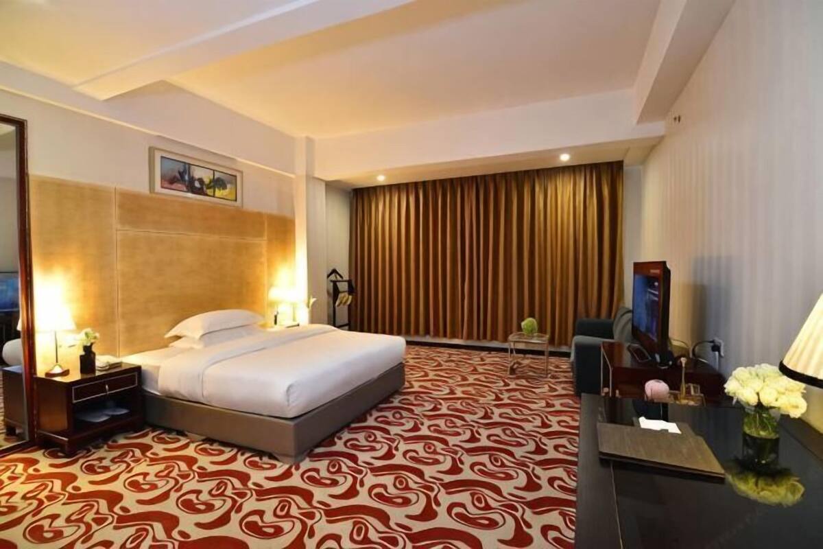 A hotel room with a bed, bedside tables, a desk, a chair, a lamp, and a large window with curtains.