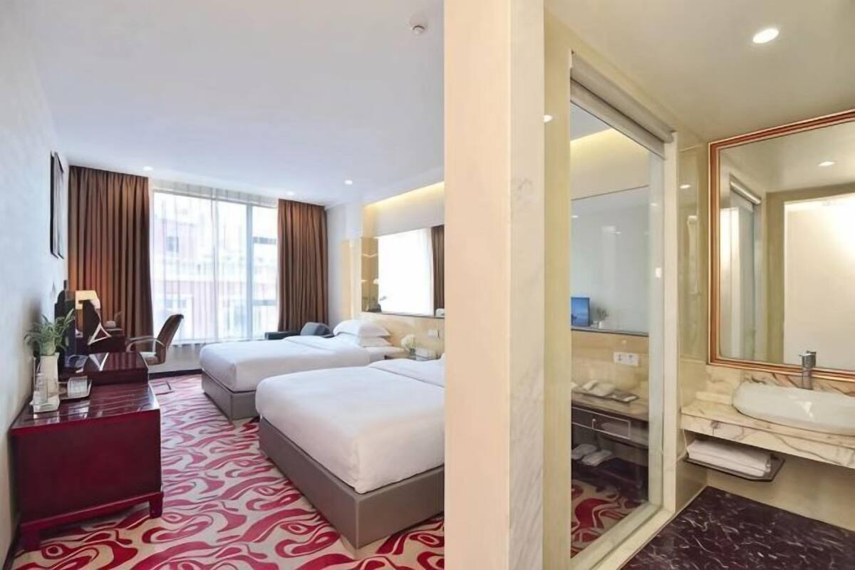 A hotel room with two beds, a desk, a chair, a large mirror, and a bathroom with a sink and mirror.