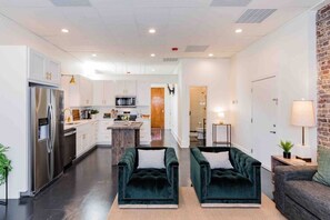 Una cocina moderna con gabinetes blancos y electrodomésticos de acero inoxidable, una isla central y un área de estar acogedora con sillas tapizadas en verde.