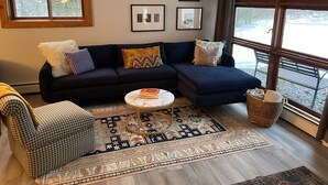 A living room with a dark blue sofa, patterned cushions, a small round coffee table, and a large window with a view of a snowy landscape.