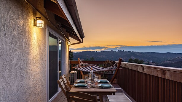 A wooden deck with a table set for two, overlooking a mountainous landscape at sunset.